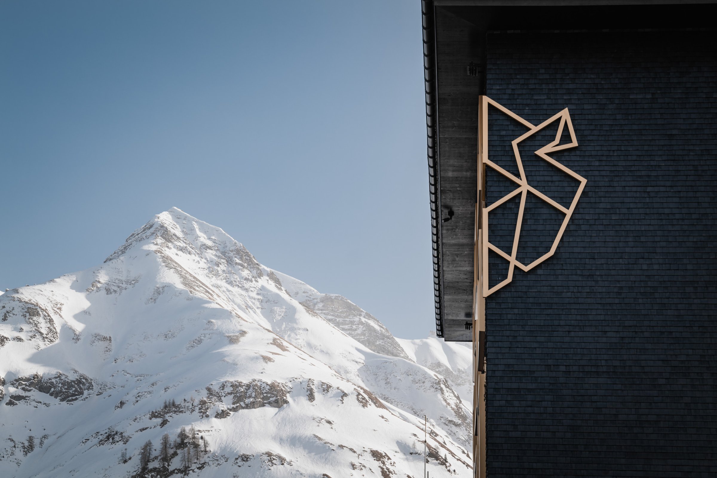 Holziges Dachsymbol an dunkler Fassade – dahinter schneebedeckte Gipfel, als hätte man das Hotel einfach zu nah ans Panorama gebaut.