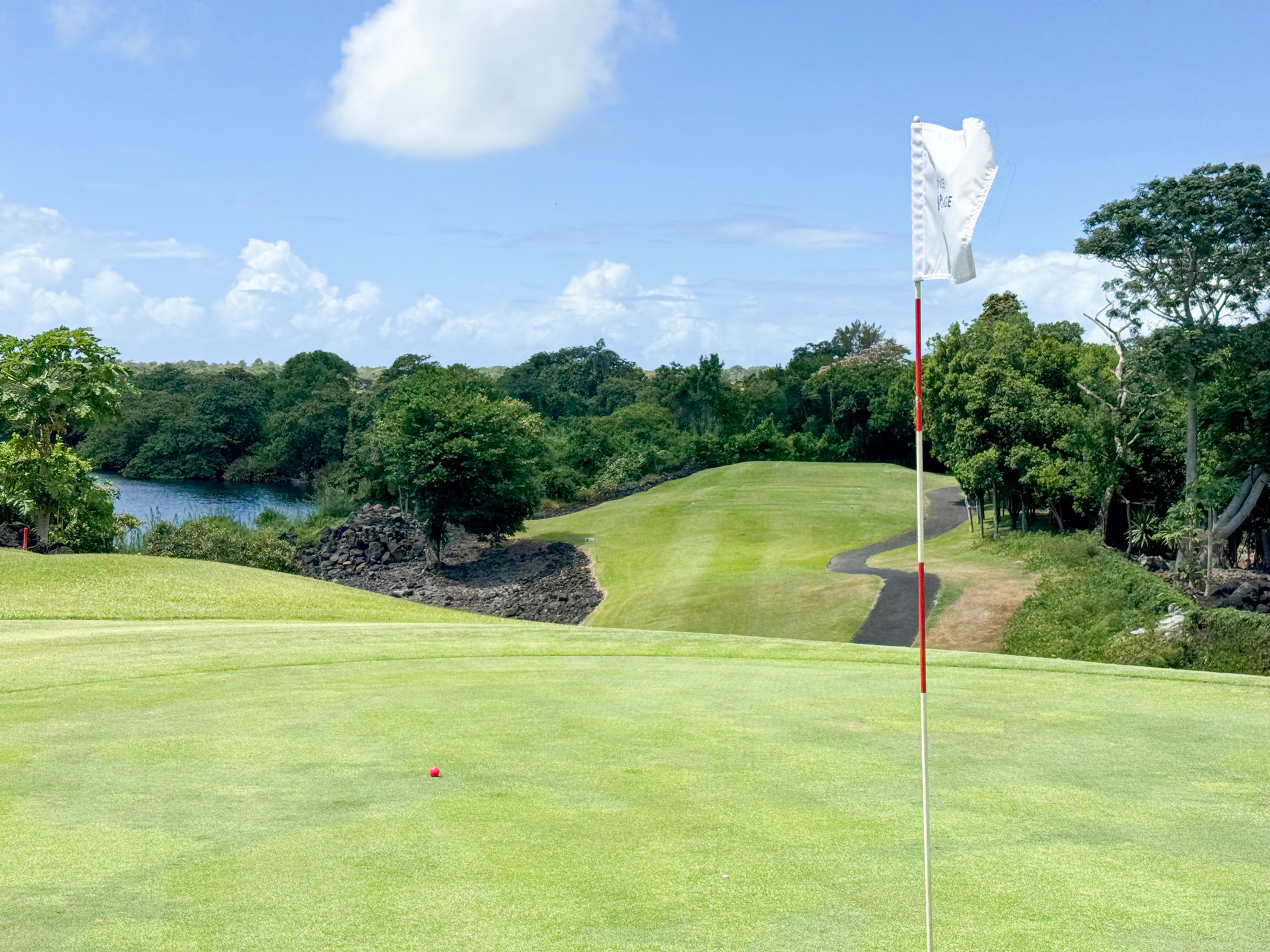 Golfgrün mit Fahne und Lavasteinen vor tropischer Landschaft unter blauem Himmel