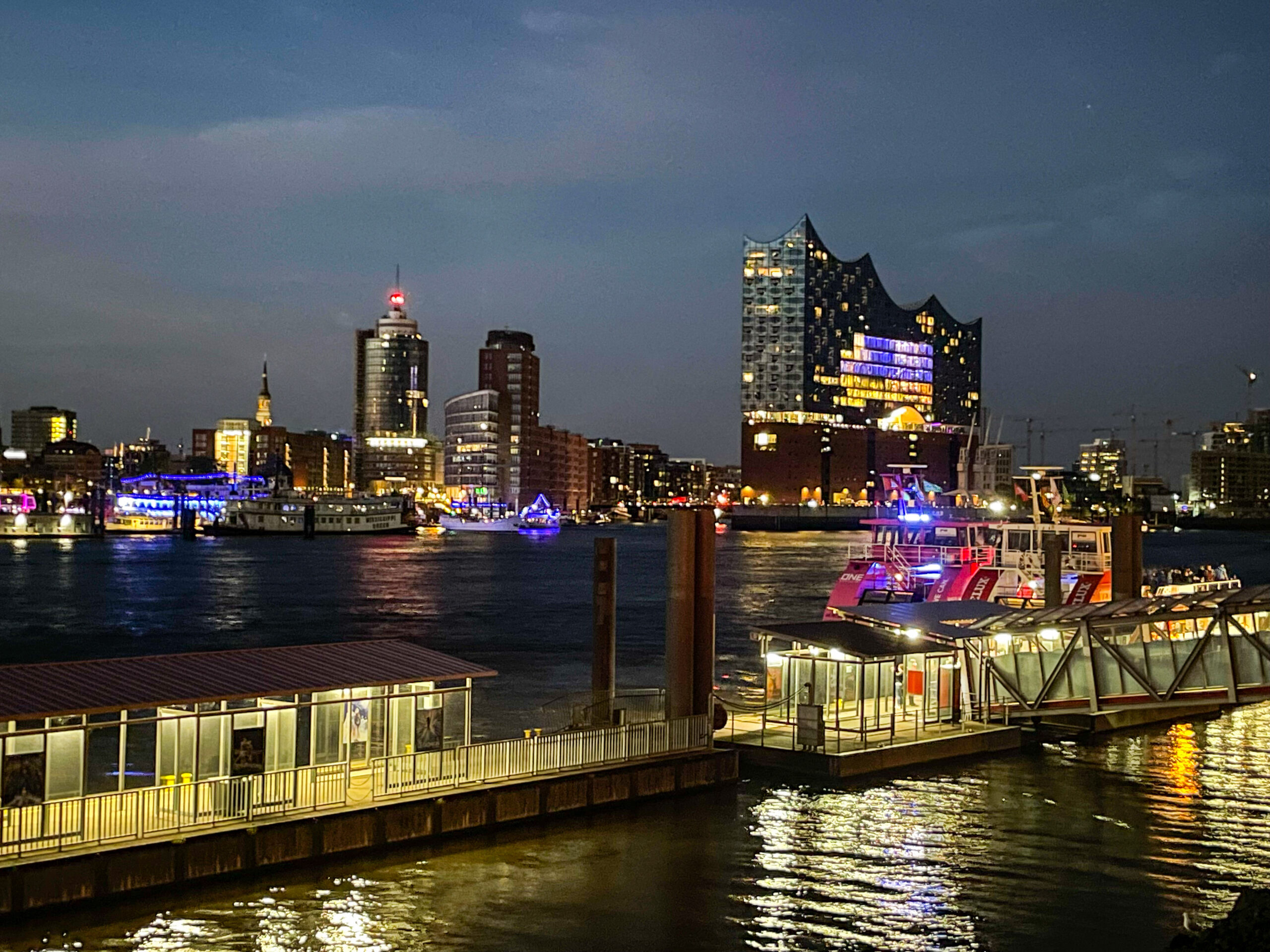Hamburgs Hafen leuchtet blau und gold in der Dämmerung – die Elbphilharmonie thront rechts wie ein imposantes Kunstwerk, während bunte Neonlichter das Wasser spiegeln und Anlegestellen im Vordergrund die typische Hafenromantik ausstrahlen.