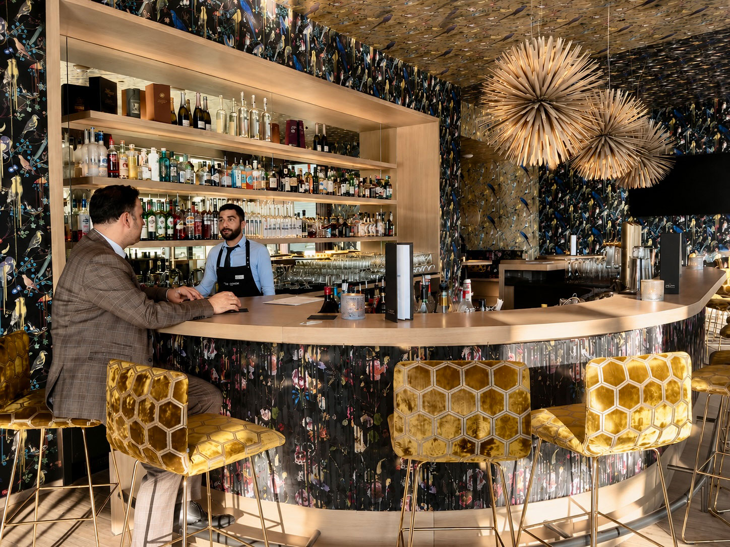 Hoteldirektor Florian spricht mit seinem Barkeeper an der Bar – während hinter ihnen 150 Flaschen warten, die definitiv weniger Aufmerksamkeit bekommen als dieser Moment.