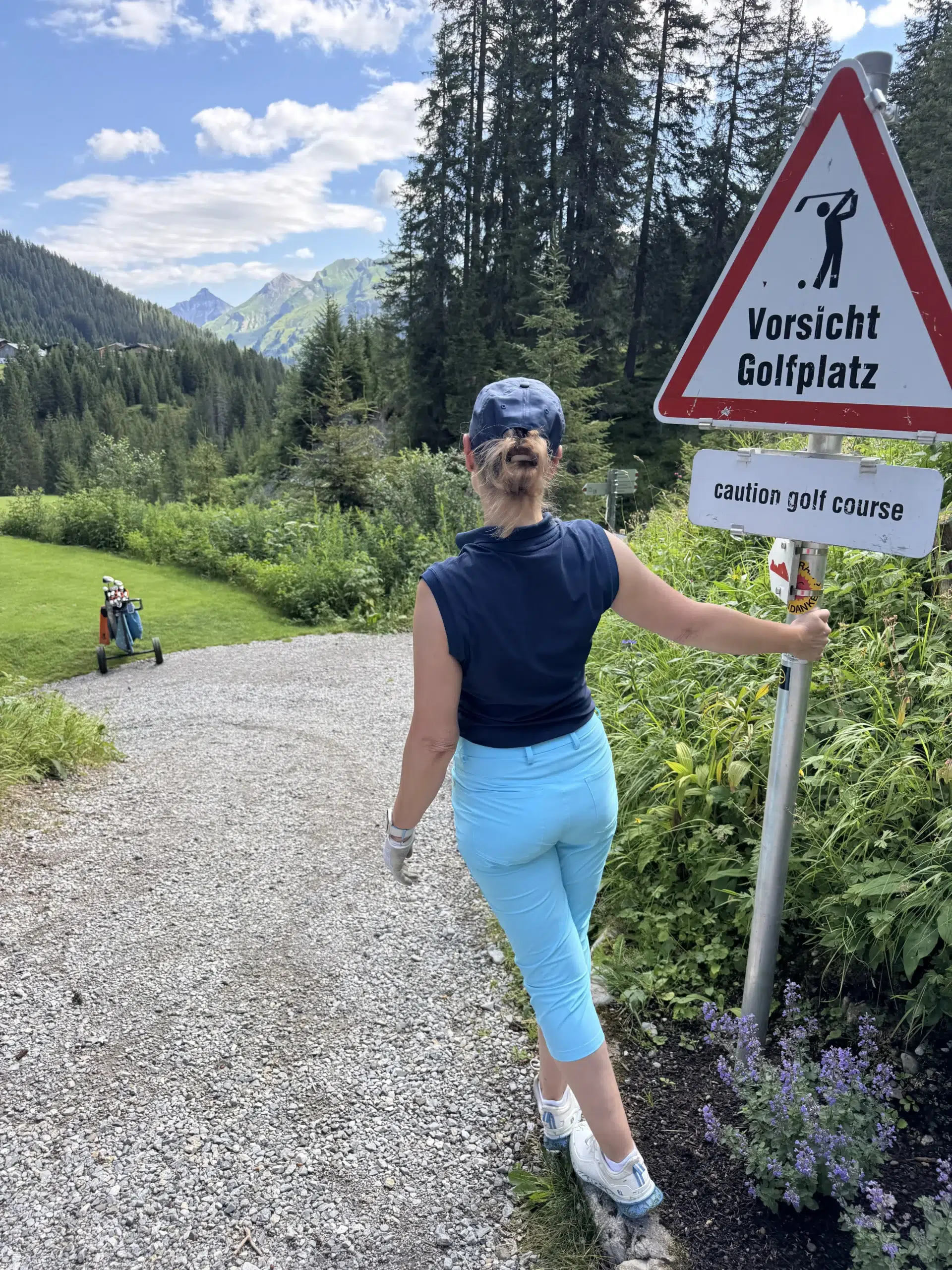Warnschild «Vorsicht Golfplatz» im Hochgebirge – dahinter Alpenwald, grüne Wiese, eine Golferin in Sportoutfit. Das ist die andere Definition von «Gefahr».