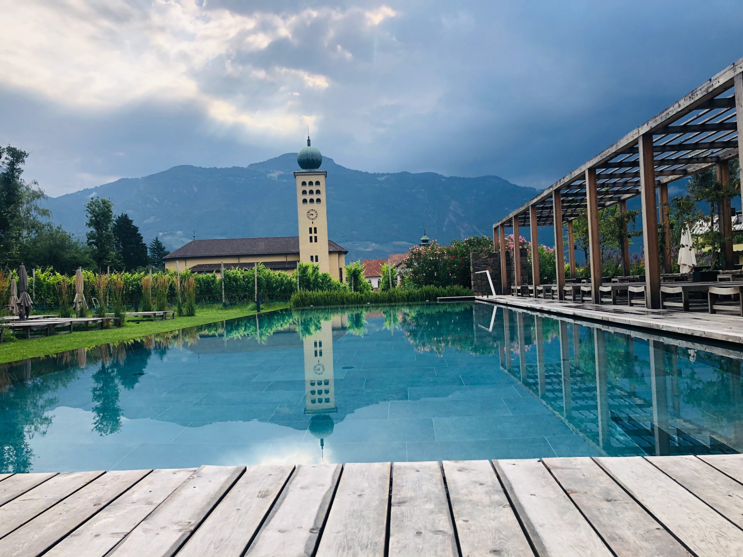 Holzsteg zum türkisen Becken – eine Kirchturmspitze spiegelt sich darin, als wolle sie sich selbst bewundern.