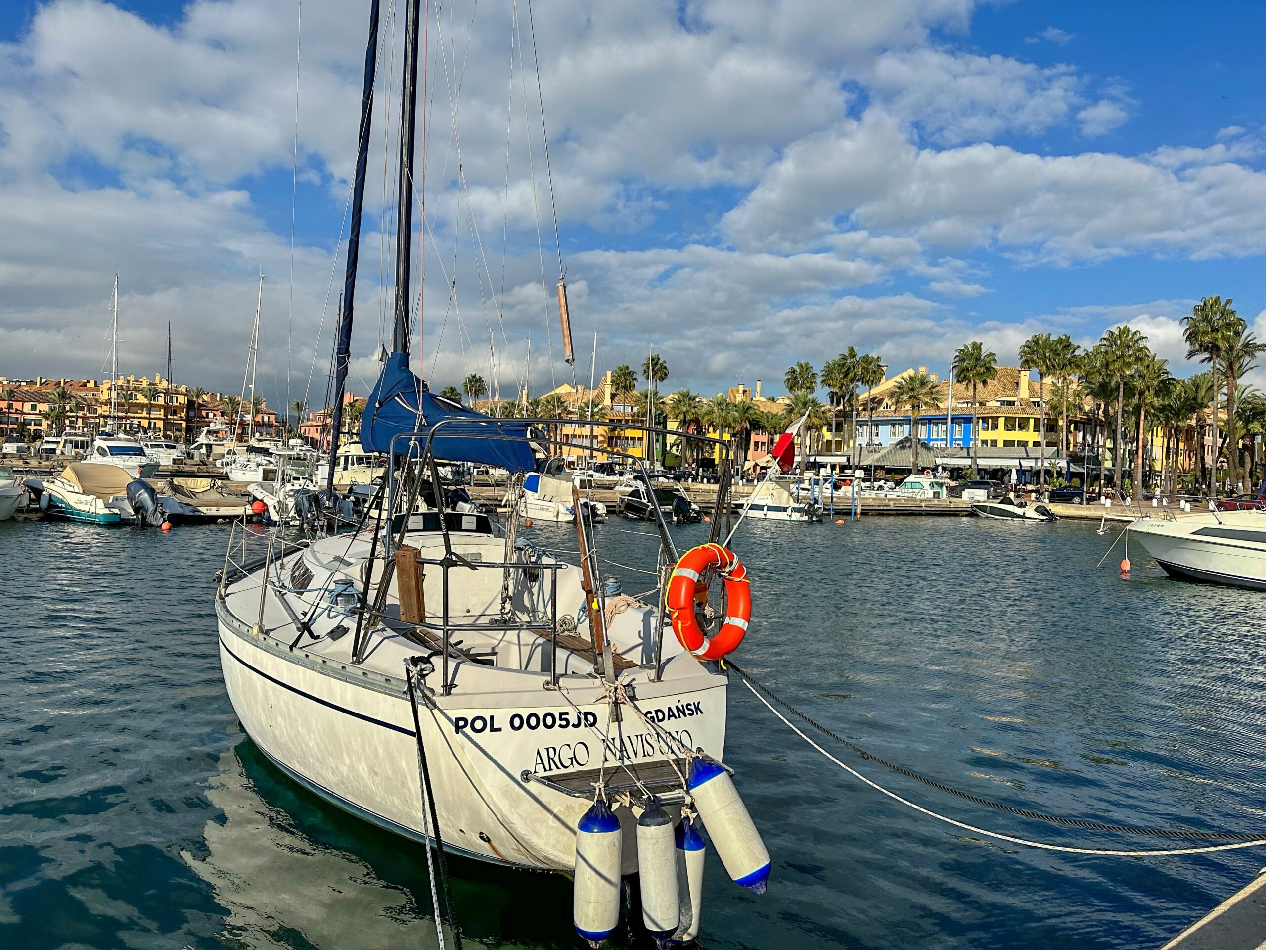 Ein weisses Segelboot mit dunkelblauem Segel liegt in der Marina – Sotogrande im Hintergrund, Palmen, Farben. Die «Argo Navissano» aus Danzig wirkt bescheiden für einen Ort wie diesen.