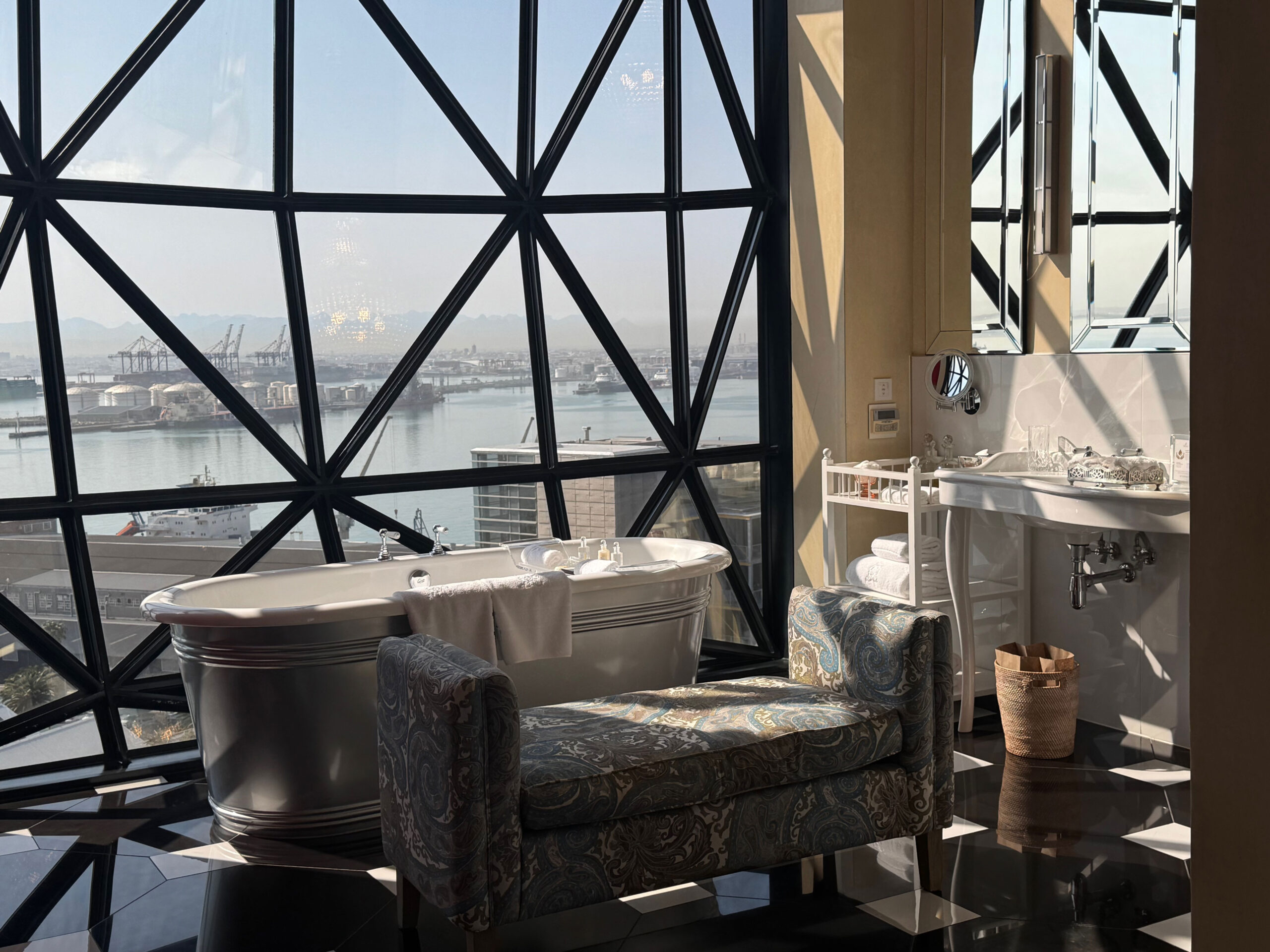 Badezimmer im The Silo Hotel mit freistehender Badewanne und grafischem Sessel – draussen die Hafenkräne von Kapstadt hinter geometrischen Fensterrahmen, als wäre die Aussicht ein Teil des Designs.